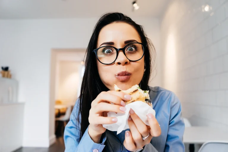 Donna con gli occhiali che mangia un panino seduta in ufficio