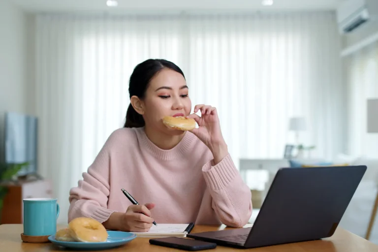 La giovane donna asiatica che mangia una ciambella guardando il monitor del notebook mentre lavora da casa